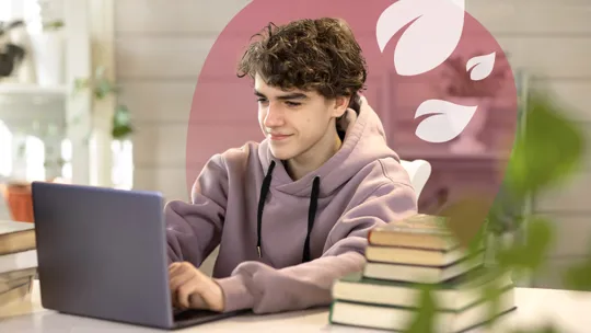 A young student with curly hair, wearing a hoodie, smiling slightly while working on a laptop at a desk with stacked books.