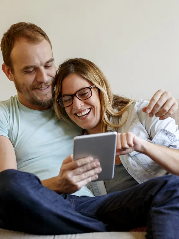 Happy couple sitting on sofa, laughing together while viewing a tablet.
