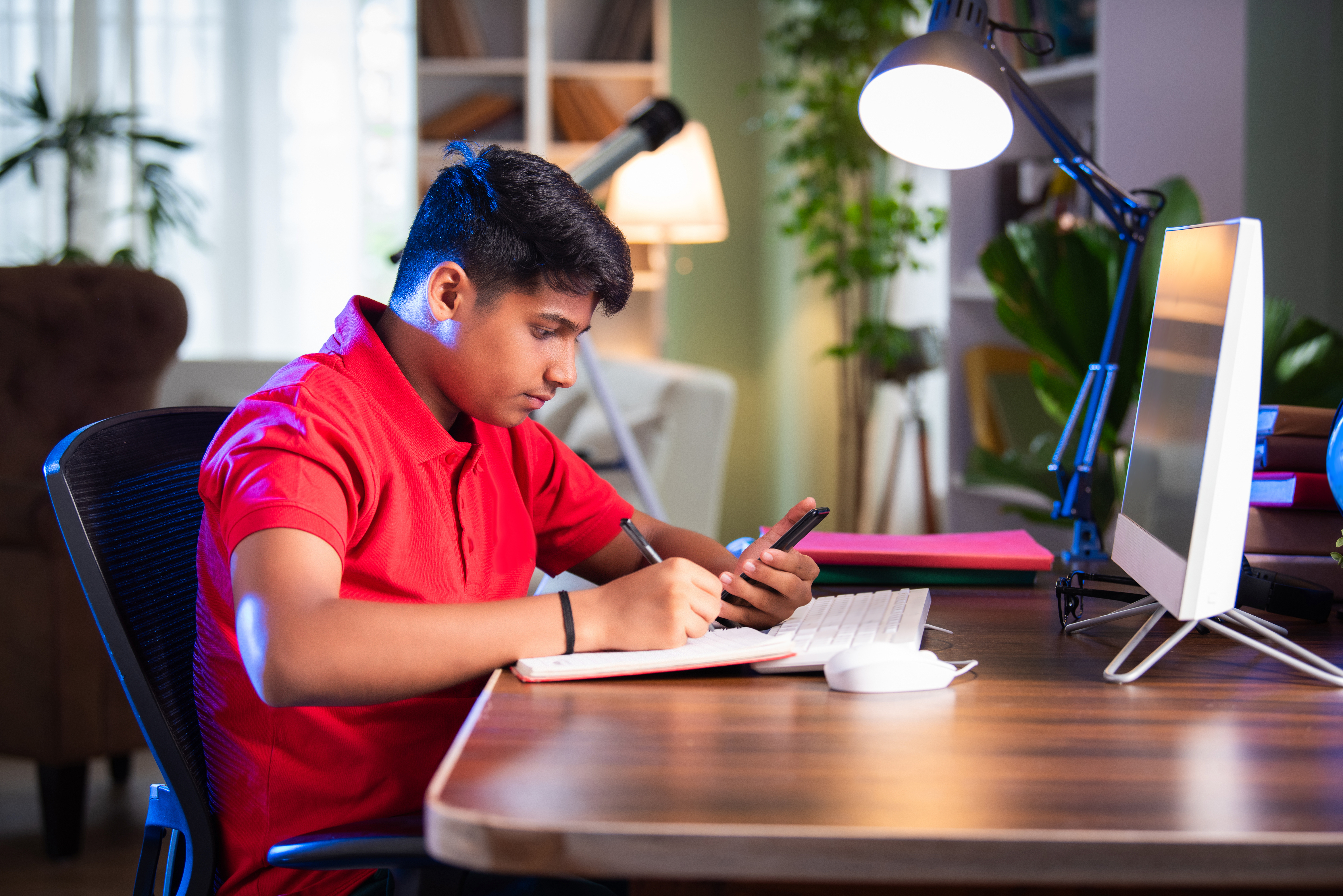 Secondary school-age student studying diligently at a desk with a computer, notebook, and smartphone.
