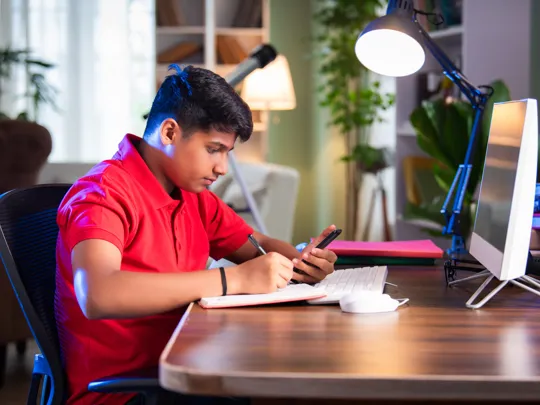Secondary school-age student studying diligently at a desk with a computer, notebook, and smartphone.