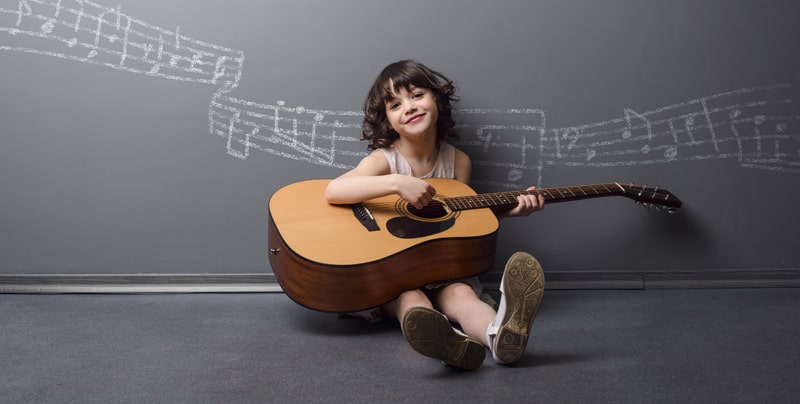 Smiling girl seated on the floor playing an acoustic guitar with chalk music notes drawn on the wall behind.