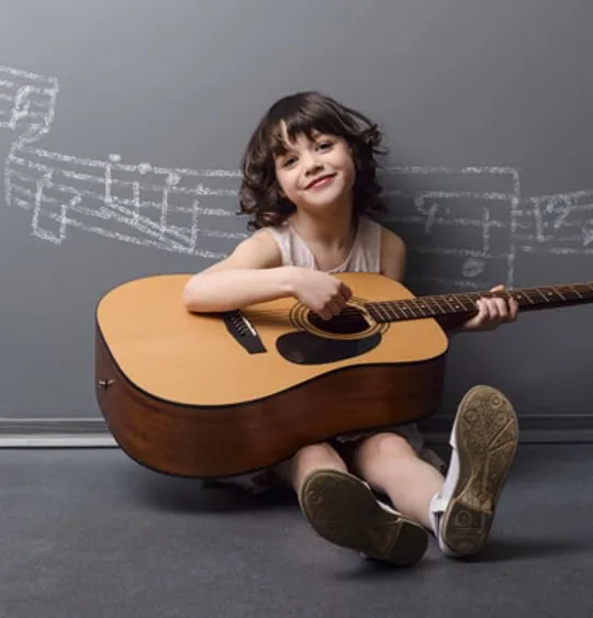 Smiling girl seated on the floor playing an acoustic guitar with chalk music notes drawn on the wall behind.