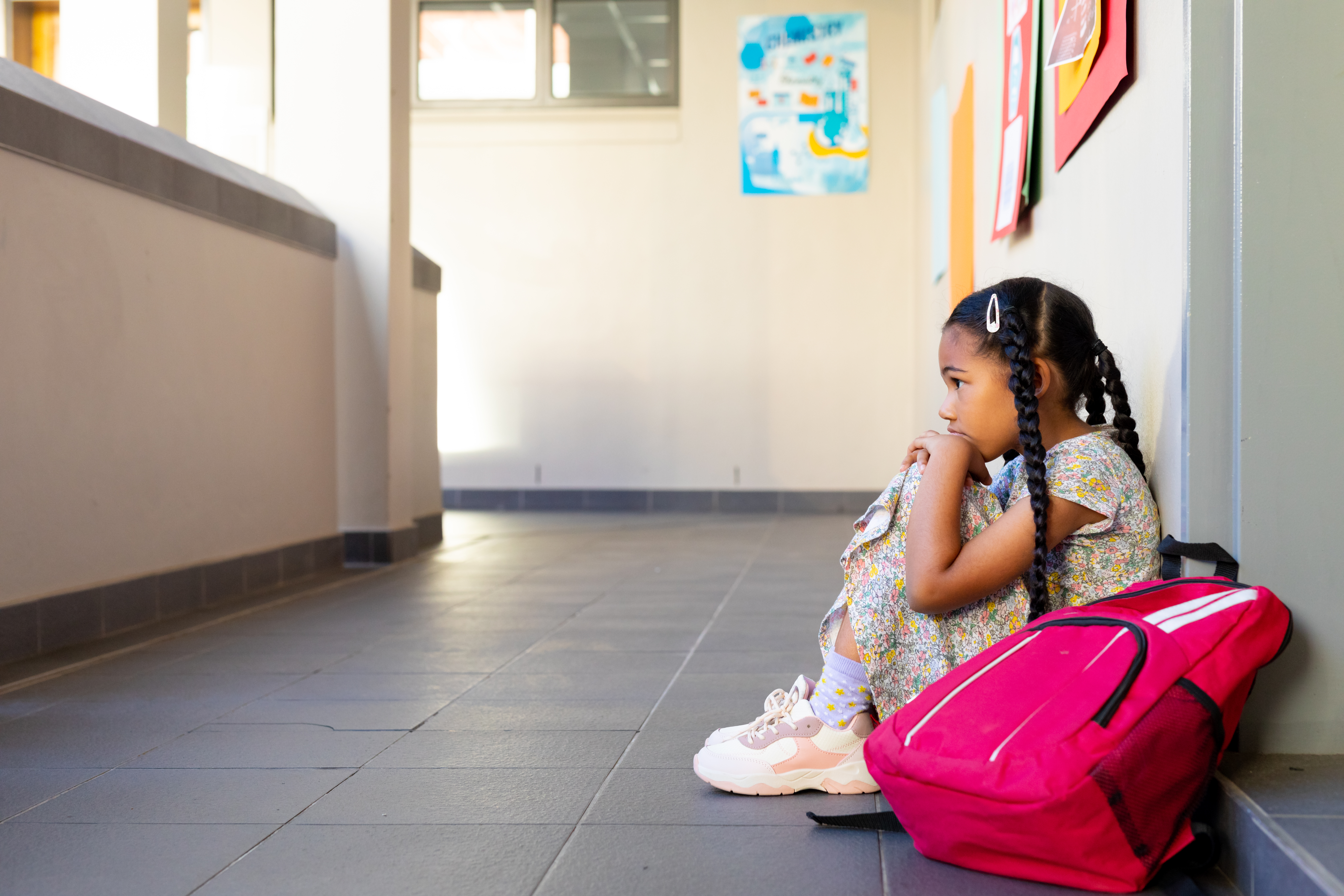 Child appearing anxious sits on the floor in a school corridor next to her bag, highlighting the topic of an event for anxious children.