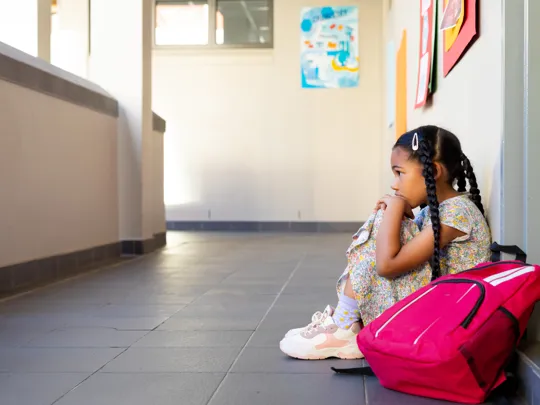 Child appearing anxious sits on the floor in a school corridor next to her bag, highlighting the topic of an event for anxious children.