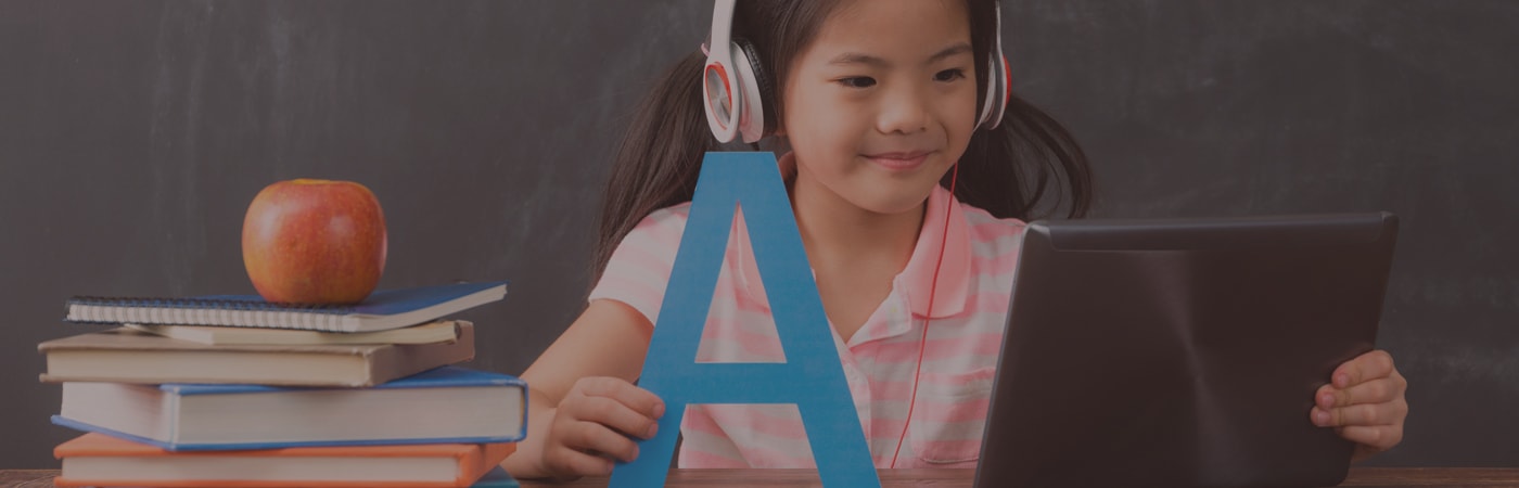 Young girl wearing headphones, learning English on a tablet with books and a large letter ‘A’ on the desk.