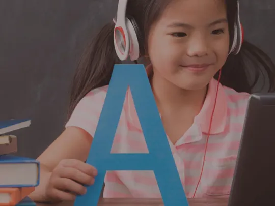 Young girl wearing headphones, learning English on a tablet with books and a large letter ‘A’ on the desk.