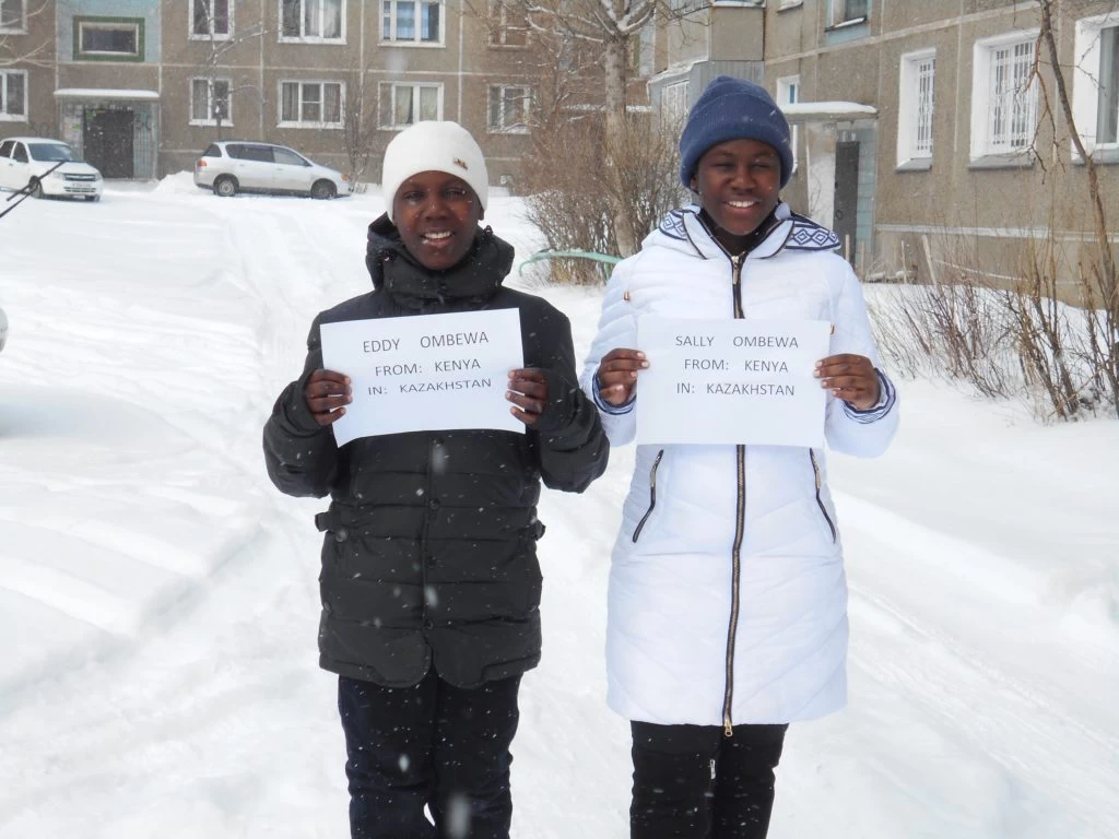 Two individuals holding signs with their names, standing in a snowy environment in front of buildings.