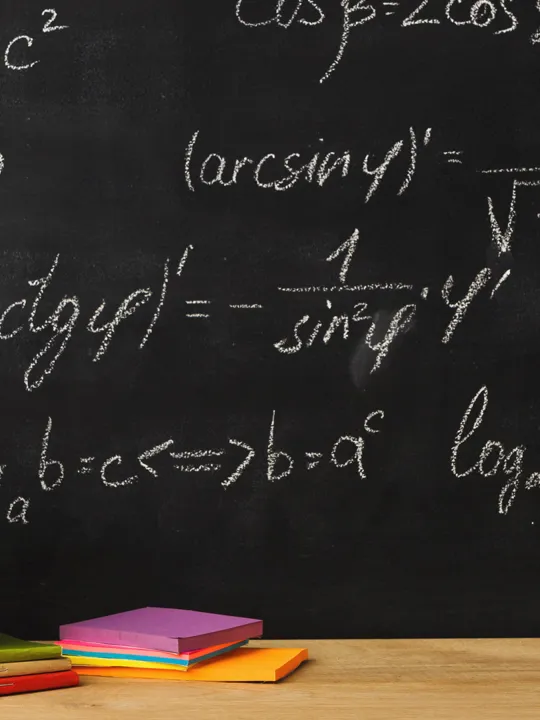 A chalkboard filled with mathematical formulas and equations is behind a wooden desk with a red apple, colorful notebooks, and a cup of writing utensils, symbolizing mathematics applications.