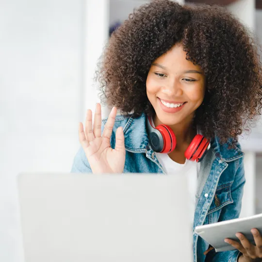 A young woman with curly hair waves during a video call on her laptop, wearing a denim jacket and red headphones around her neck, representing online learning.