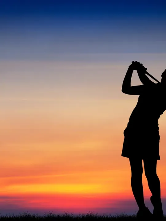 A silhouette of a golfer completing a swing against a colourful sunset sky.
