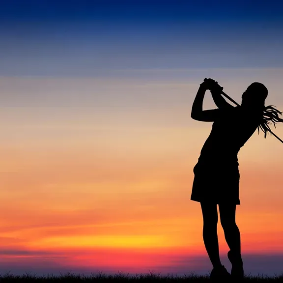 A silhouette of a golfer completing a swing against a colourful sunset sky.