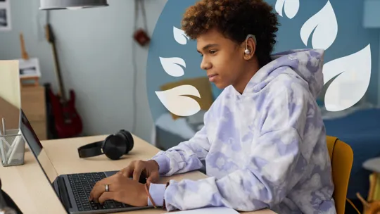 Focused boy wearing a hearing aid, studying at his desk with a laptop and notebook.