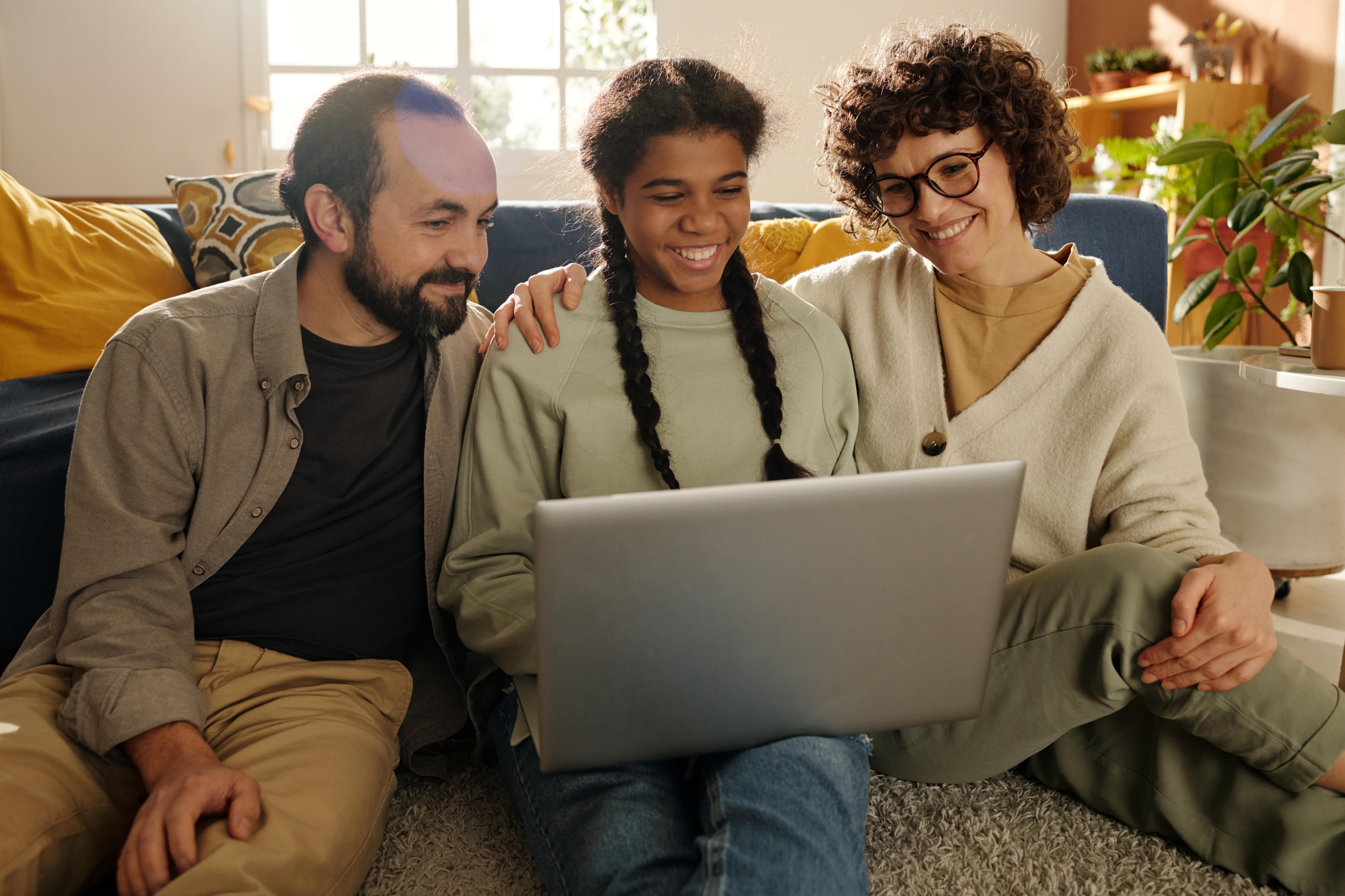 A smiling mother, father, and their teenage daughter sit together on the living room floor, happily watching something on a laptop.