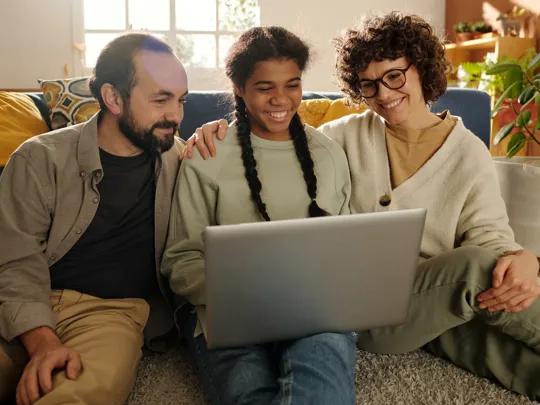 A smiling mother, father, and their teenage daughter sit together on the living room floor, happily watching something on a laptop.