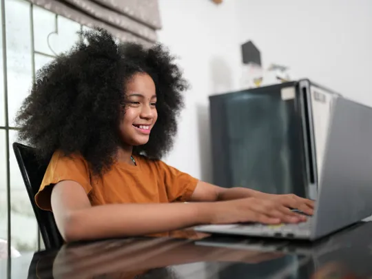 Young girl smiling as she types on a laptop at a kitchen table.