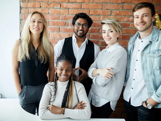 Group of five young professionals smiling together in a modern office with brick walls.
