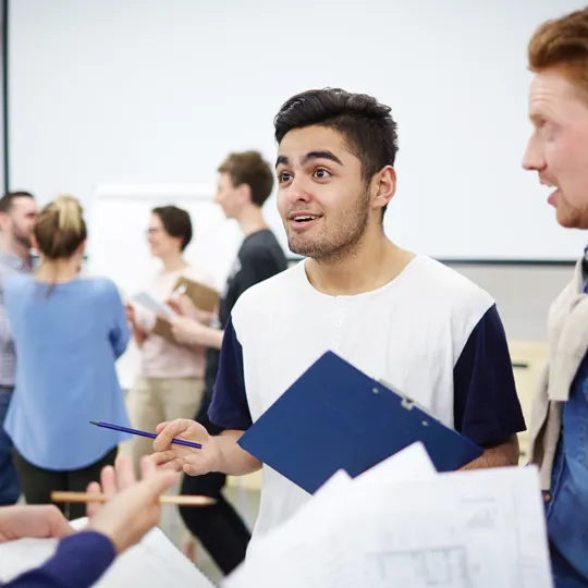 A young man holding a clipboard smiles and gestures with a pencil while engaging in an excited discussion with a small group of classmates in a modern classroom setting.