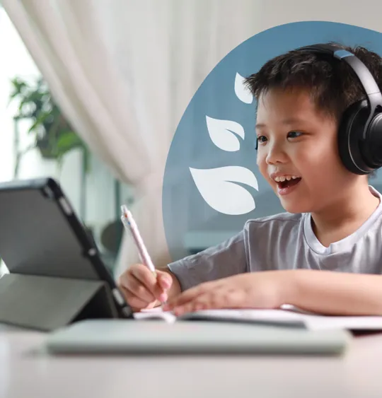 A young student wearing headphones, smiling and writing in a notebook while looking at a tablet, indicating active online learning.