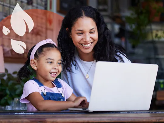 An adult and a young girl smiling and looking at a laptop together, appearing to be in a cafe setting.