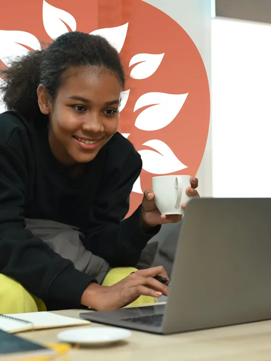 Smiling student with a mug in hand using a laptop at home, seated on a sofa with notebooks on the table.