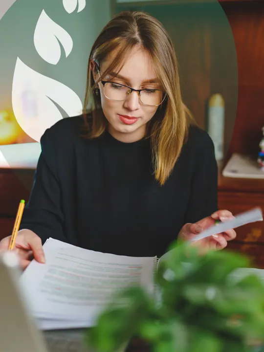 A young woman with glasses holding papers and writing, seated at a desk with a laptop and a plant, with a green leafy design in the background.