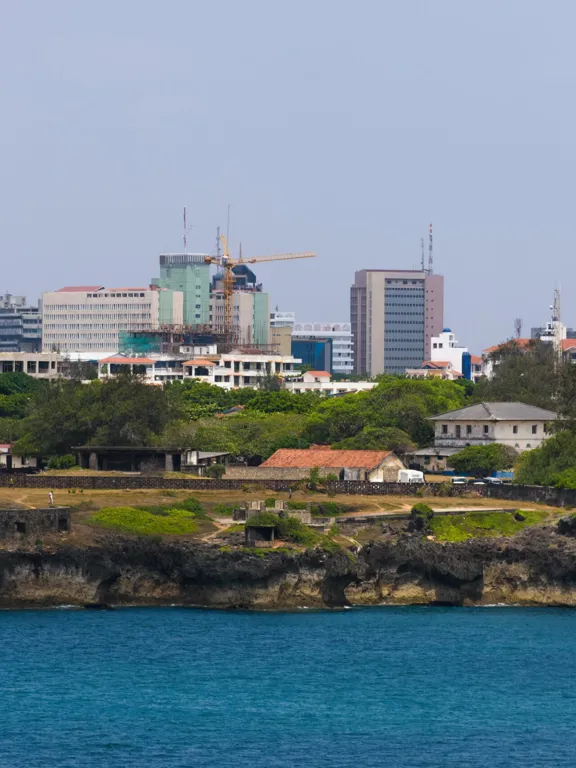 Cityscape of Mombasa viewed from across the water, with modern buildings and coastal vegetation.