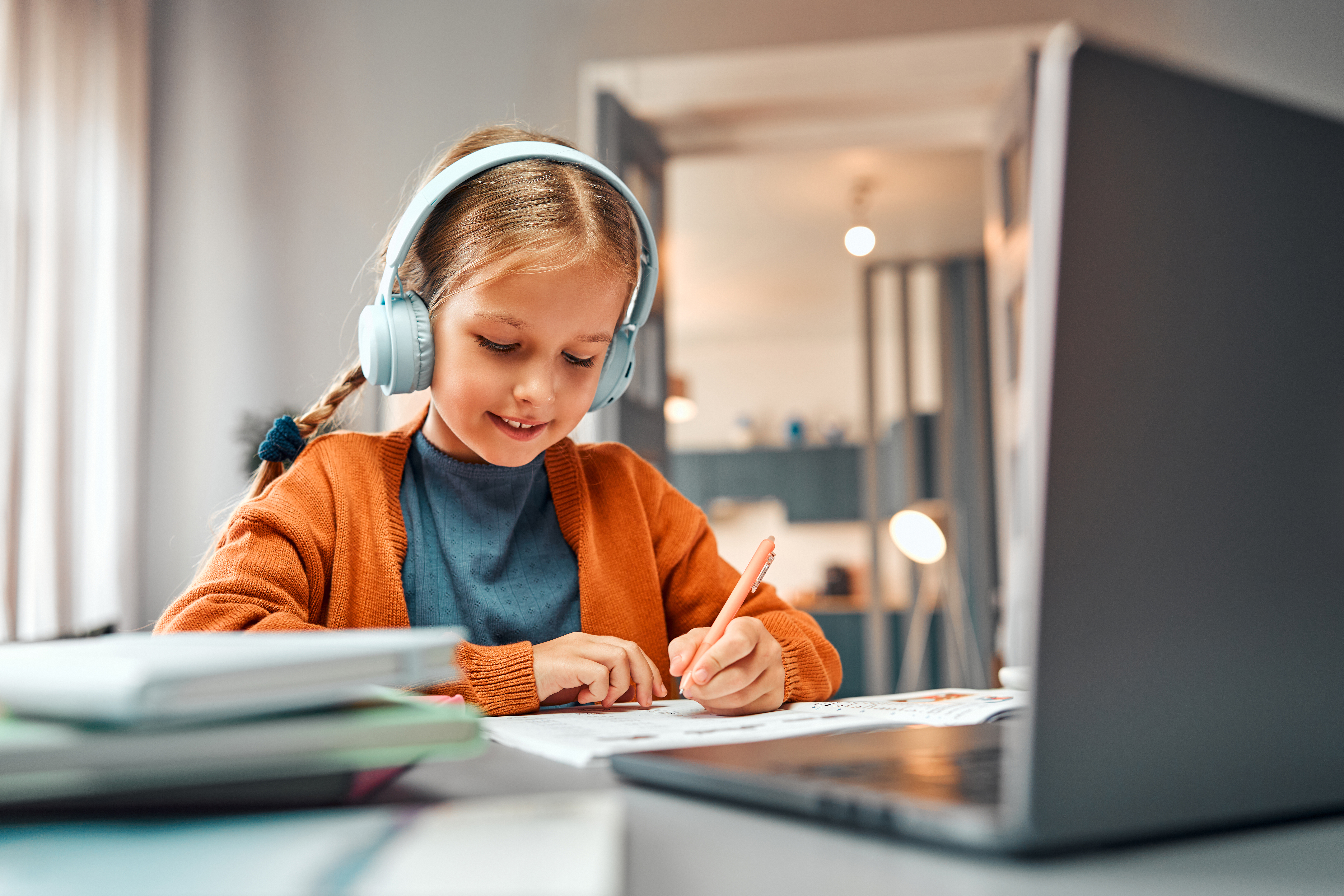 A young girl wearing headphones and an orange cardigan is focused on her studies, writing in a workbook while using a laptop for online learning at her desk.