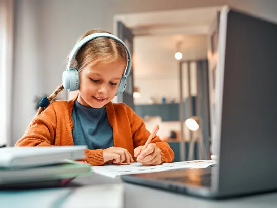 A young girl wearing headphones and an orange cardigan is focused on her studies, writing in a workbook while using a laptop for online learning at her desk.