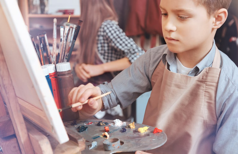 Student concentrating on a painting at an easel, mixing colours on a palette during an art lesson