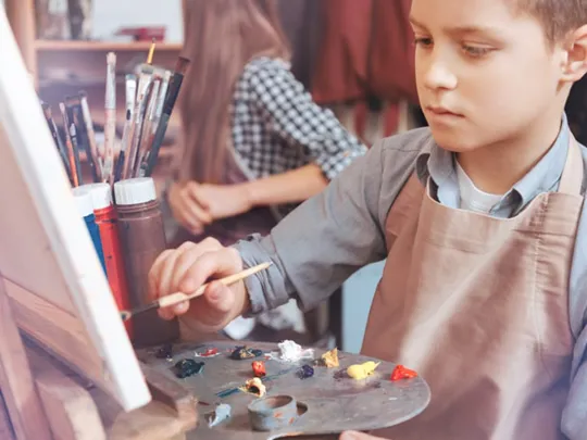 Student concentrating on a painting at an easel, mixing colours on a palette during an art lesson