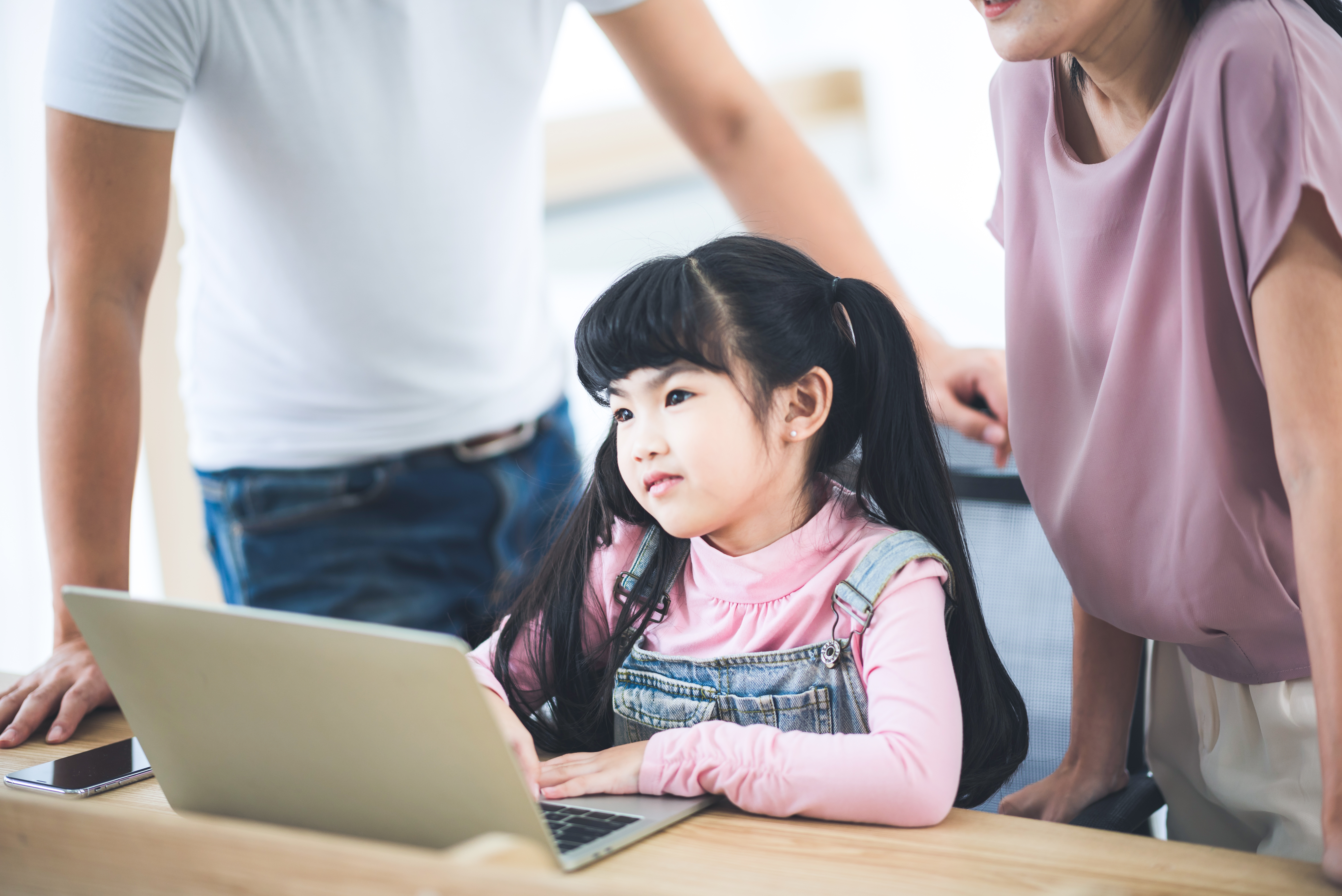 Young girl with long hair working on a laptop, supported by her parents in a bright room