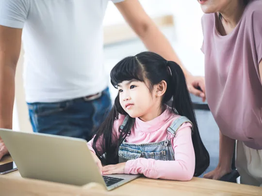 Young girl with long hair working on a laptop, supported by her parents in a bright room