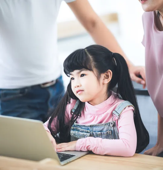 Young girl with long hair working on a laptop, supported by her parents in a bright room