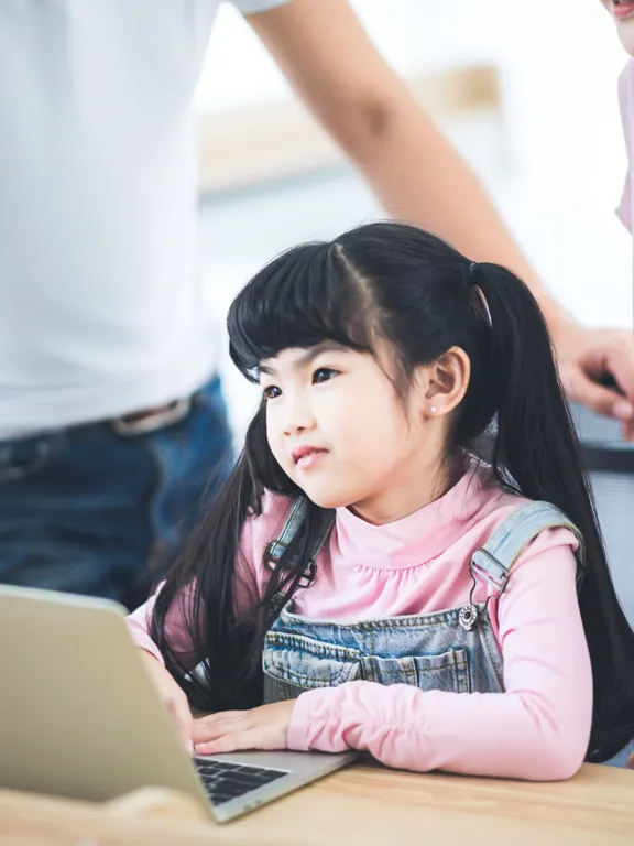 Young girl with long hair working on a laptop, supported by her parents in a bright room