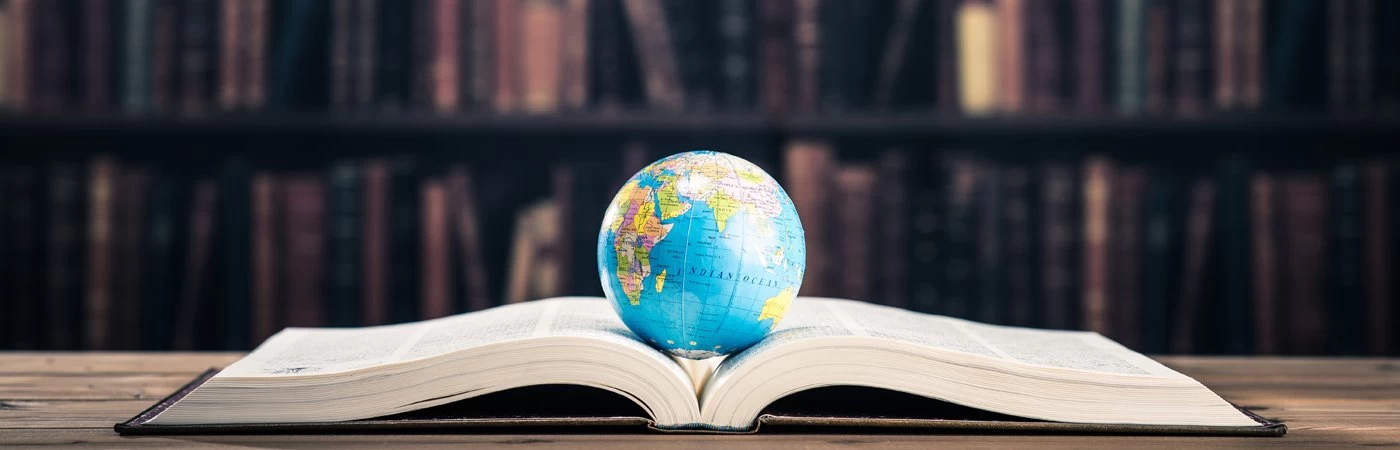 A large book within a library open at the middle with a globe laid on top of it. 
