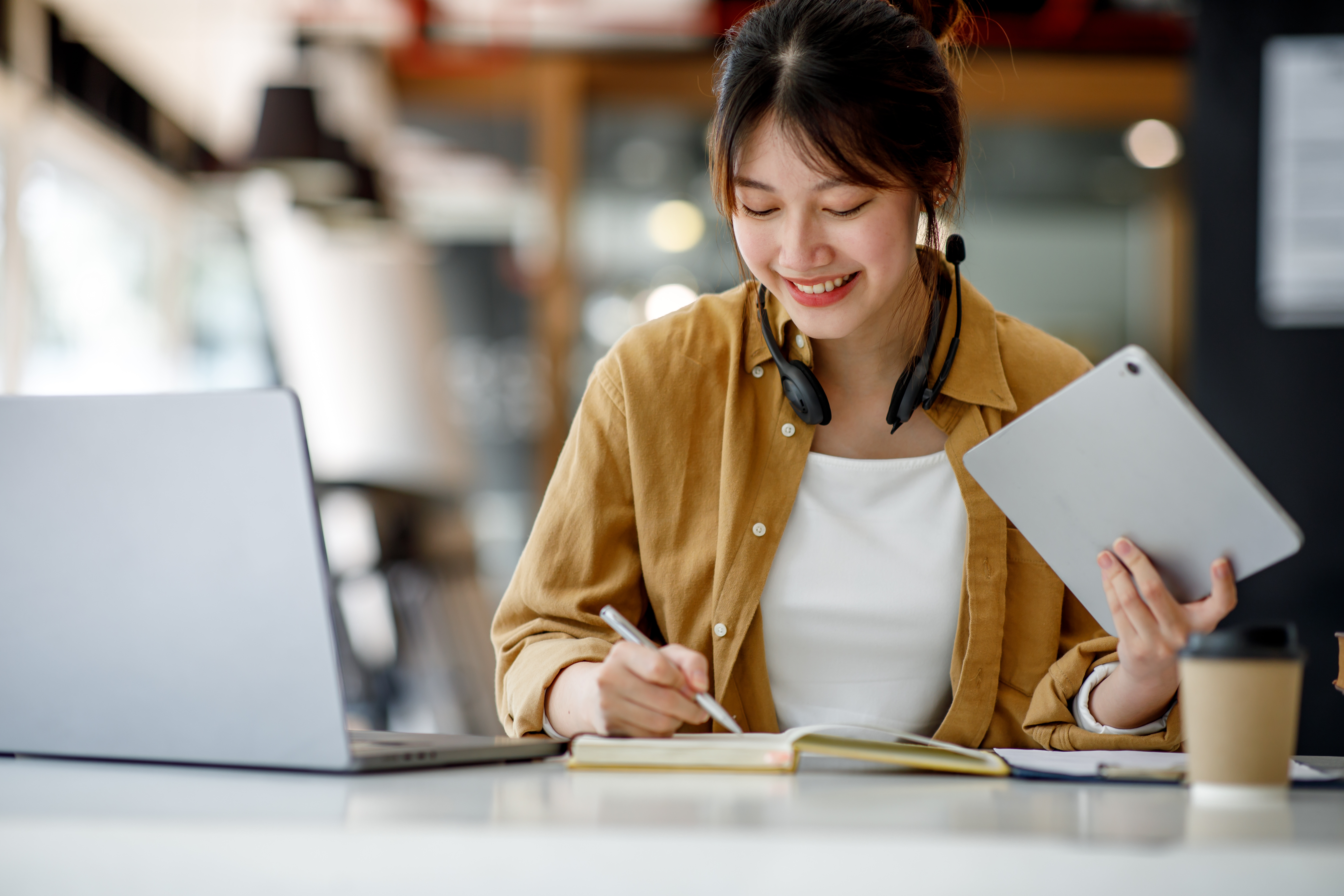 A young woman smiles while writing in a notebook at a desk, holding a tablet in her other hand, with a laptop and coffee cup nearby, diligently preparing for A Level mock exams.