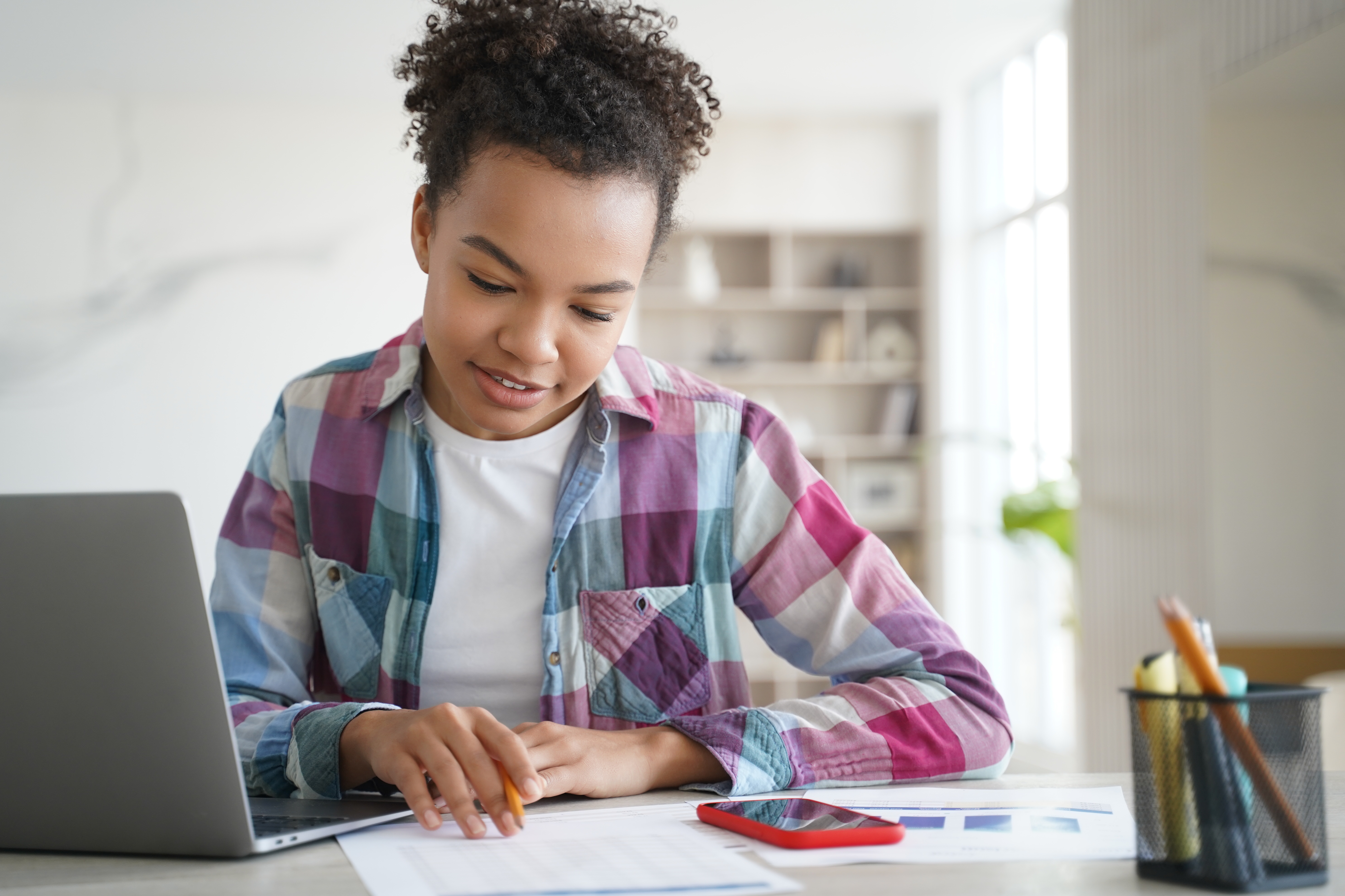 Focused student working with laptop and documents in a bright home setting.
