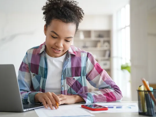 Focused student working with laptop and documents in a bright home setting.