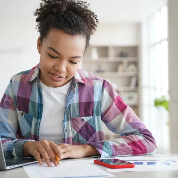 Focused student working with laptop and documents in a bright home setting.