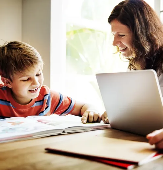 A mum helping her young son homeschool. They are both sat smiling at a desk with a laptop and a textbook.