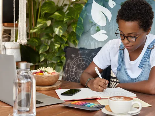 Student writing in a notebook beside a laptop and a bowl of fresh fruit.