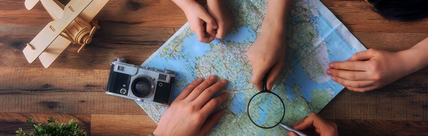 Hands of children and adults pointing at a paper map with a magnifying glass, camera and toy plane nearby
