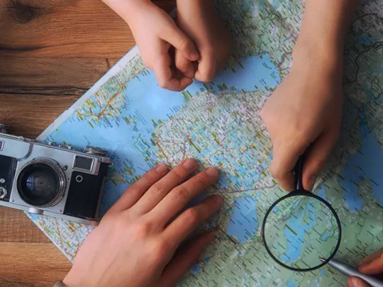 Hands of children and adults pointing at a paper map with a magnifying glass, camera and toy plane nearby