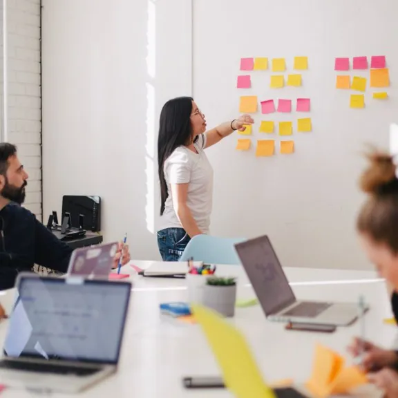 A group of people in an office or classroom setting. A woman is standing at a whiteboard covered in colorful sticky notes, pointing to one of them while speaking. Other individuals are seated around a table with laptops and notebooks, looking at the whiteboard.