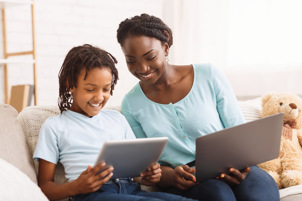 A homeschooling mother and son sitting on a sofa. The mother is holding a laptop and the son is holding a tablet; they are both looking at the tablet and smiling.