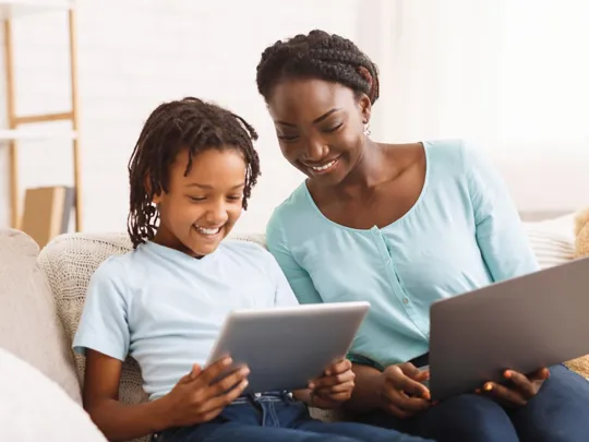 A homeschooling mother and son sitting on a sofa. The mother is holding a laptop and the son is holding a tablet; they are both looking at the tablet and smiling.