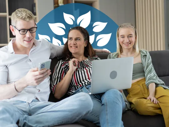 Three people sitting on a sofa smiling while looking at a laptop and phone together