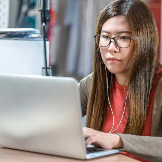 A young woman wearing glasses and earbuds intently works on a silver laptop at a desk, representing focused online learning or remote work.