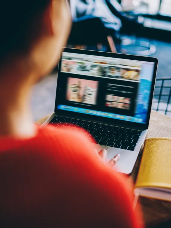 A person in a red shirt working on a laptop displaying a website photo grid, with a yellow notebook nearby, set in a trendy cafe.