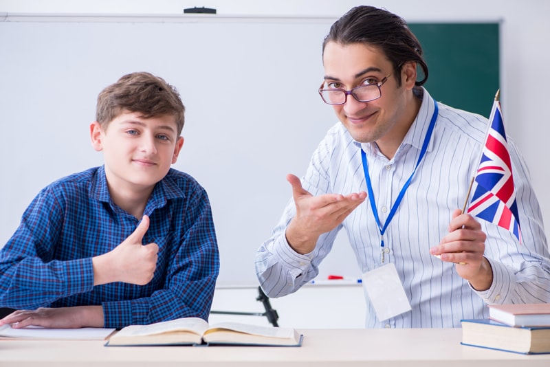 Smiling English teacher holding a UK flag next to a student giving a thumbs up in a classroom.